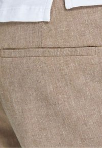 Pantalons en lin beige avec une texture subtile, dotés d'une poche passepoilée à l'arrière. Le tissu a une apparence en fibres naturelles.