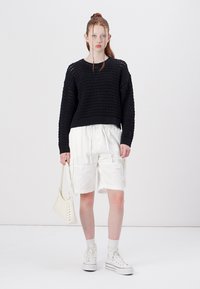 Un pull noir en tricot avec une coupe ample, un short cargo blanc et des baskets montantes blanches. Portant un petit sac à dos blanc.