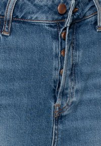 Světle modré džíny s vysokým pasem, obsahující pět bronzových knoflíků a viditelné švy, texturovaná tkanina s opotřebovaným vzhledem.