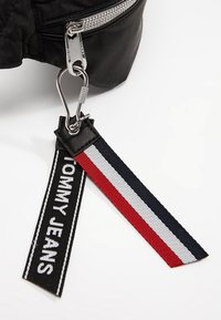 Svart tygväska med en silverdragkedja och en avtagbar rem som har en svart etikett med "TOMMY JEANS" och röda, vita och blå linjer.