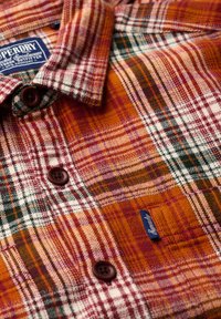 Camisa de franela a cuadros en rojo y naranja con detalles en verde, que presenta botones marrones y una etiqueta azul. Patrón texturizado con cuadros variados.