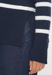 Ναυτική ριγωτή μπλούζα με λευκές οριζόντιες ρίγες, ριμπ ωστόσο, πλευρικό σκίσιμο και φαρδιά μανίκια, συνδυασμένη με σκούρο μπλε τζιν.