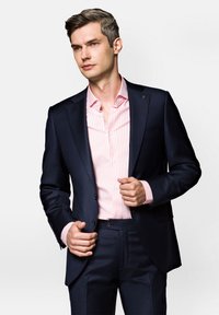 Ciemnoniebieska marynarka w zestawie z jasnoróżową koszulą w paski, o dopasowanym kroju i detalach z guzikami, noszona z dopasowanymi spodniami.