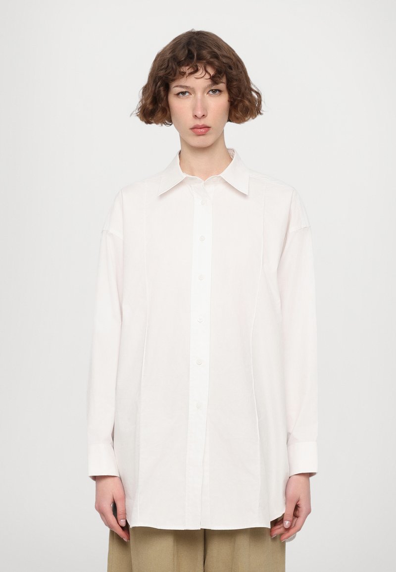 Camicia bianca con colletto, maniche lunghe e vestibilità rilassata. Presenta dettagli di cucitura sottili e una texture liscia.