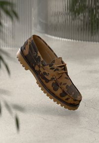 Mocassim com padrão de camuflagem em castanho e preto, com sola de borracha, sobre um chão de concreto claro e rachado, e um fundo metálico desfocado.