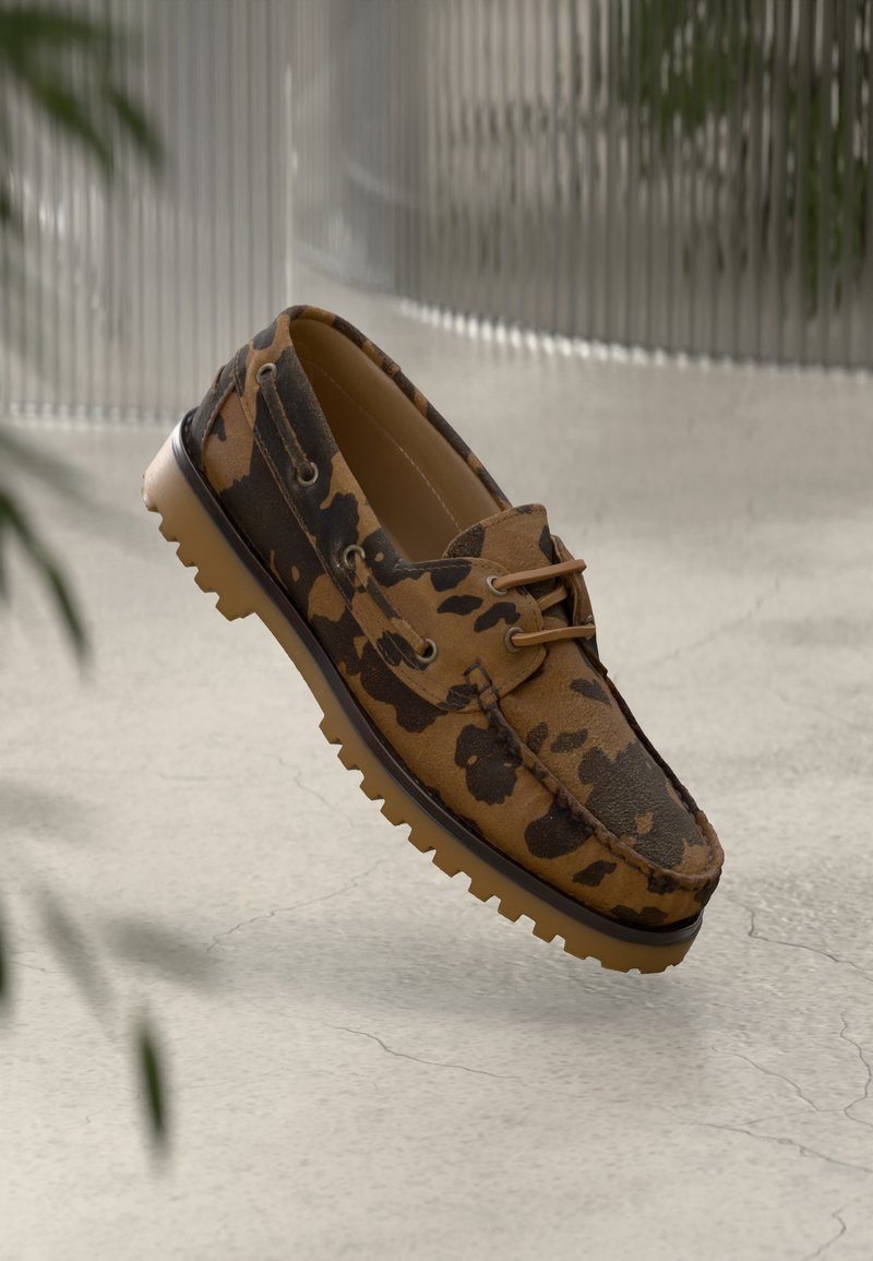 Mocassim com padrão de camuflagem em castanho e preto, com sola de borracha, sobre um chão de concreto claro e rachado, e um fundo metálico desfocado.