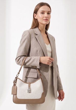 Femme portant un blazer beige et une robe crème tient un sac à main beige et blanc avec une fermeture à verrou argenté sur l'épaule.