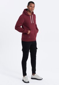 Felpa bordeaux con cappuccio, cordini bianchi, tasca frontale e polsini a costine; abbinata a pantaloni cargo neri e sneakers grigie.