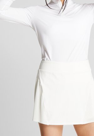 Frau trägt ein weißes, langärmeliges Oberteil und einen weißen, hoch taillierten, plissierten Rock, steht mit erhobenen Armen vor einem schlichten Hintergrund.