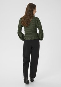 Frau mit langen braunen Haaren, die eine grüne geraffte Bluse, schwarze Hosen und schwarze High Heels trägt, steht mit dem Rücken zu einem weißen Hintergrund.