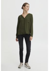 Blusa verde oliva a maniche lunghe con scollo a V, tasca sul petto e dettaglio logo circolare, abbinata a jeans skinny neri e sneaker grigie.