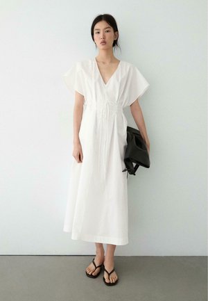 Kobieta w białej sukience midi z dekoltem w serek i szerokimi rękawami, trzymająca czarną torebkę i nosząca czarne sandały, stojąca na tle jednolitej ściany.