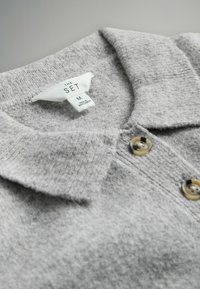 Sivá polo majica z mehkimi vlakni, gumbom za zapiranje in zloženo ovratnikom. Na označbi za nego je navedena velikost M in blagovna znamka "THE SET."