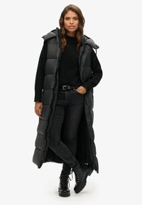 Langer, ärmelloser Daunenwesten in Schwarz, mit Kapuze, seitlichen Schlitzen und gestepptem Muster. Getragen über einem schwarzen Pullover und dunklen Jeans, mit schwarzen Stiefeln.