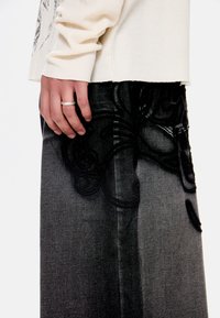 Το χέρι ενός ατόμου με ασημένιο δαχτυλίδι ακουμπάει πάνω σε μαύρο και γκρι παντελόνι με ανάγλυφη σ κούρα λουλουδάτη κεντητική, φοράει κρεμ υφασμάτινη μπλούζα με μακριά μανίκια.