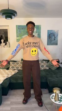 Fiatal férfi mosolyogva, kinyújtott karokkal, bézs "Szerencsés vagyok! Látom a szellemeket" pulóvert és sötét nadrágot viselve egy díszített nappaliban.