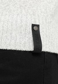 Materiale di maglione bianco lavorato a maglia con una trama a coste, con un'etichetta in tessuto nero con ferramenta argentata con la scritta "INDICODE." Abbinato a tessuto nero.