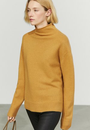 Donna con capelli biondi corti che indossa un maglione a collo alto giallo senape e pantaloni neri, che tiene le maniglie di una borsa marrone.