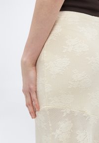 Χέρι γυναίκας ακουμπισμένο στη μέση, φοράει κρέμ φούστα από δαντέλα με λουλουδάτο σχέδιο και σκούρο κορυφαίο ρούχο, πάνω σε απλό ανοιχτό φόντο.