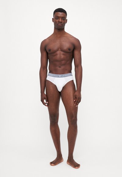 Witte katoenen slips met een grijze merkband, met een aansluitende pasvorm en een minimalistisch ontwerp, gedragen door een model die blote voeten heeft.