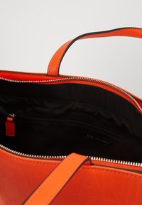 Pomarańczowa torba na ramię z wytłoczonym logo, wyposażona w czarne wnętrze i zamknięcie na zamek błyskawiczny. Zawiera małą wewnętrzną kieszeń do organizacji.