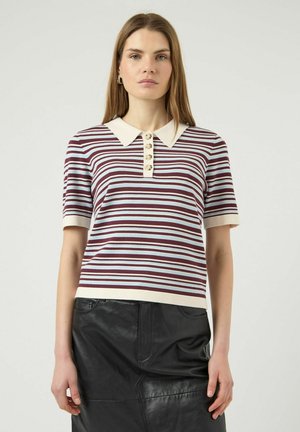 Vrouw draagt een shirt met korte mouwen, een kraag, in bordeauxrood en wit gestreept, en een zwarte leren rok, staand tegen een effen achtergrond.