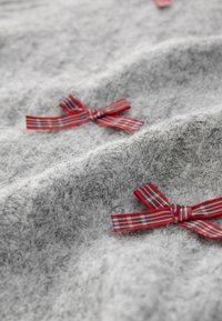 Tissu gris texturé avec des nœuds en ruban écossais rouge et vert cousus sur la surface, créant un effet superposé.