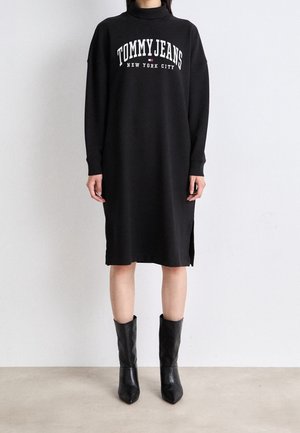 Osoba ubrana w czarną sukienkę midi z długim rękawem z napisem "TOMMY JEANS New York City", w połączeniu z czarnymi skórzanymi botkami do połowy łydki.