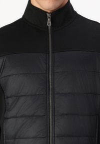 Mustlane jakk, millel on tõmblukuga kinnitus, sujuva ülemise osa ja tikitud, polsterdatud alumise osaga. Kõrge krae, neutraalsed toonid.