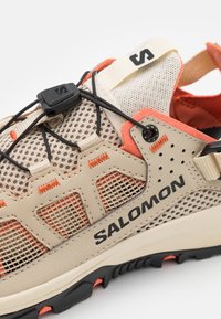 Chaussure de plein air en mesh beige avec des accents orange vif. Comprend un système de laçage sécurisé, une semelle en caoutchouc texturée et le logo "SALOMON".
