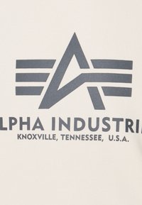 Tessuto color crema con un design grafico grigio scuro. Il logo include un triangolo astratto e strisce orizzontali con il testo "Alpha Industries".