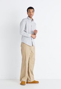 Ljusblå skjorta med knapp-up och en subtil mönstring, matchad med beige kordflorsbyxor och bruna slip-in skor. Modellen står mot en vit bakgrund.