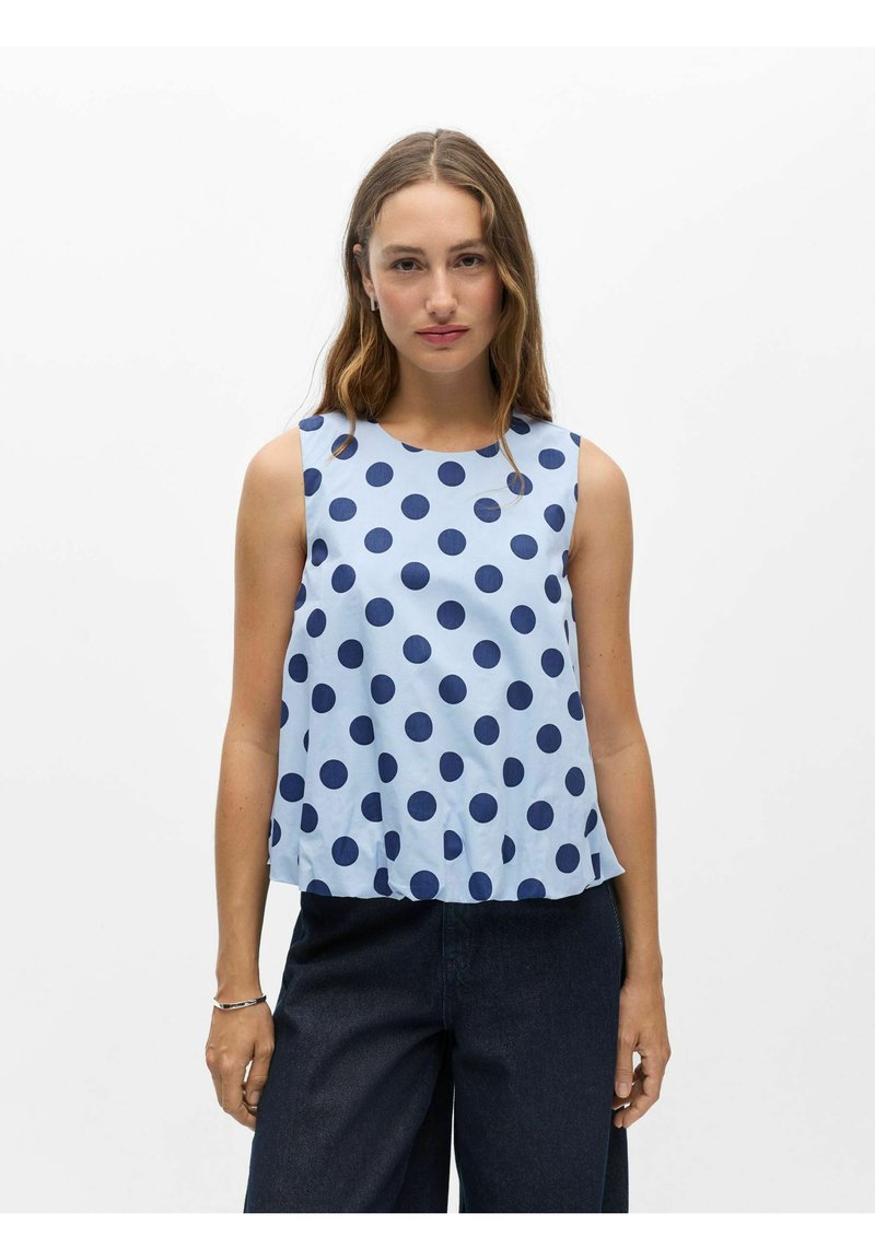 Femme aux longs cheveux portant un haut sans manches bleu clair à gros pois bleu foncé et un pantalon foncé sur un fond blanc uni.