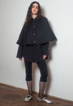 Młoda kobieta z długimi kręconymi włosami nosi czarną kurtkę w stylu peleryny, krótkie legginsy, przezroczyste rajstopy i srebrne baletki wewnątrz pomieszczenia.