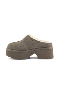 Bruine suède klomp met een fluffy witte voering, lage platformzool en een klittenbandsluiting voor aanpassing. Geribbelde rubberen zool voor grip.