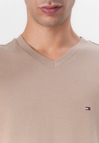 T-shirt beige à col en V en tissu doux avec une coupe ajustée. Présente un petit logo avec des accents rouge, blanc et bleu sur la poitrine.