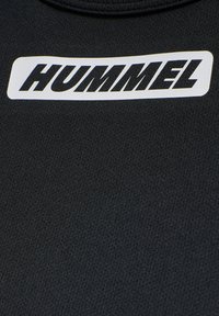 Schwarzer strukturierter Stoff mit weißem rechteckigem Logo, das den fetten schwarzen Schriftzug "HUMMEL" im Brustbereich zeigt.