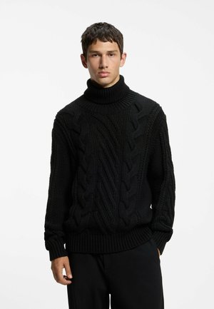 Jeune homme portant un pull à col roulé épais en maille torsadée noire et un pantalon noir, debout devant un fond blanc uni.