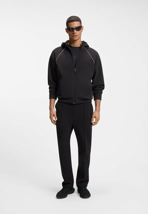 Mann trägt schwarzen Kapuzenpullover mit Reißverschluss, passende schwarze Hose, schwarze Turnschuhe und dunkle Sonnenbrille, steht vor einem schlichten weißen Hintergrund.