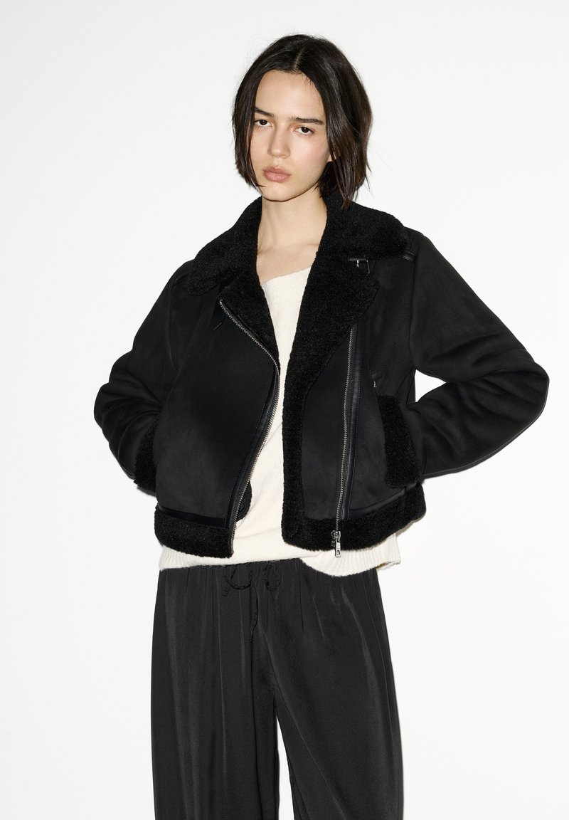 Giacca nera in shearling con chiusura frontale con zip, ampio colletto e texture accogliente. Indossata sopra una maglia chiara e abbinata a pantaloni neri.