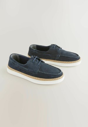 Scarpe slip-on in suede blu navy con suole in gomma bianca e un dettaglio di corda di juta lungo il bordo, presentano un semplice design con lacci.