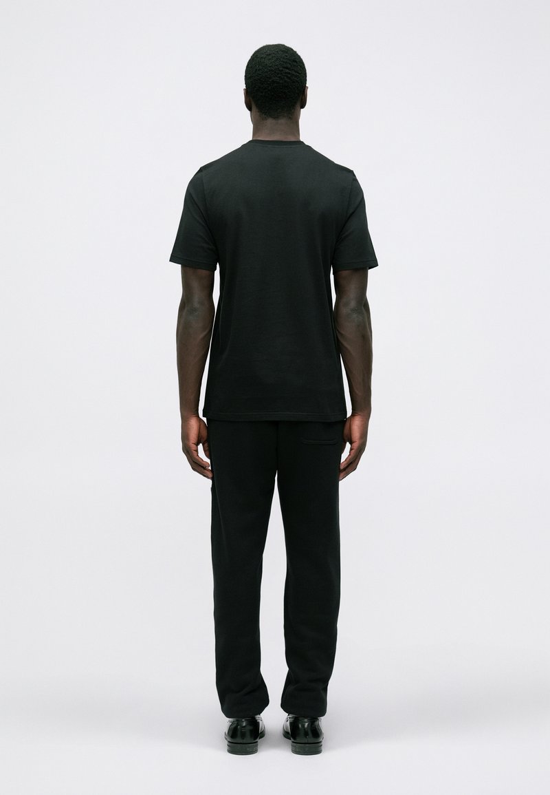 Mann, der mit dem Rücken steht und ein schwarzes kurzärmeliges T-Shirt, schwarze Hosen und schwarze Schuhe gegen einen schlichten weißen Hintergrund trägt.