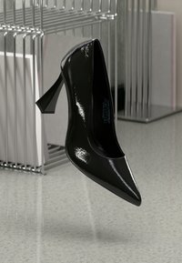 Pantof cu toc înalt din piele patent neagră, cu vârf ascuțit și tocul stiletto curbat și unghiular, suspendat deasupra unui podea netedă, de culoare deschisă.