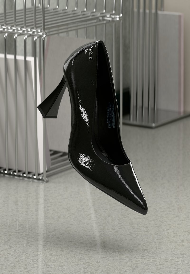 Pantof cu toc înalt din piele patent neagră, cu vârf ascuțit și tocul stiletto curbat și unghiular, suspendat deasupra unui podea netedă, de culoare deschisă.