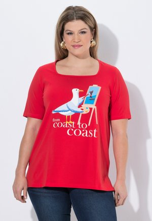 Kobieta w czerwonej koszuli z obrazem mewy malującą łódź na sztaludze, z napisem "od wybrzeża do wybrzeża".