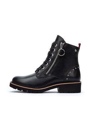 Botas negras de cuero hasta el tobillo con tacón apilado, cremallera lateral y diseño de cordones. Presentan tachuelas de metal y una tira de agarre roja en contraste.