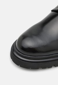 Black leather boot with a rounded toe, smooth surface, textured black sole, and a subtle contrasting seam detail at the base.