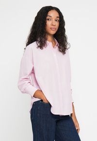 Chemise boutonnée rose clair en tissu lisse, dotée d'un col classique, de manches longues et d'une coupe décontractée. Portée avec un jean foncé.