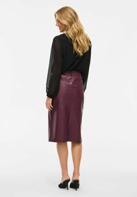 Žena s dlhými blond vlasmi, oblečená v čiernej priesvitnej blúzke, fialovej koženej sukni po kolená a čiernych lodičkách na vysokom opätku, stojí na bielom pozadí.