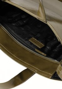 Innenseite einer olivgrünen Tasche aus Kunstleder mit einem Reißverschlussfach, ausgekleidet mit einem schwarzen Musterstoff und einem kleinen Markenetikett.